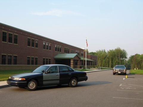 1991 Ford LTD Crown Victoria - Two sedans are parked in a lot outside a brick building with an American flag flying.