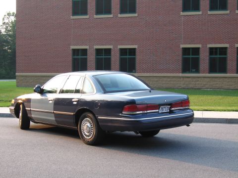 1991 Ford LTD Crown Victoria - A dark blue sedan with a two-tone front fender is parked on asphalt in front of a brick building.