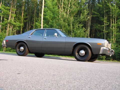 1991 Ford LTD Crown Victoria - Side profile of a gray sedan, likely a 1977 Plymouth Gran Fury, parked on an asphalt road with a forest in the background.