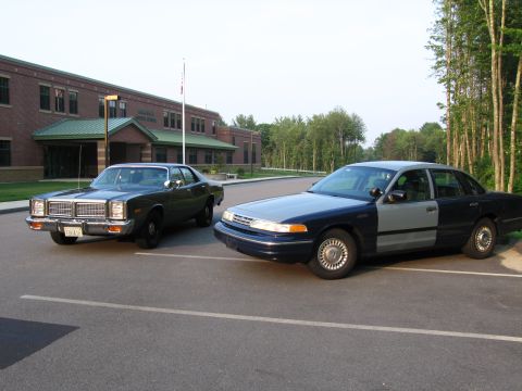 1991 Ford LTD Crown Victoria - Two sedans are parked in a school parking lot. The dark green sedan is older, while the blue and gray sedan appears to be a later model.