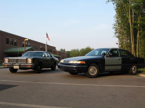 1991 Ford LTD Crown Victoria - A black and gray Ford Crown Victoria is parked next to an older dark sedan in front of a brick building.