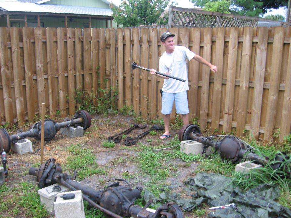 Man holding a driveshaft among rusty truck axles on cinder blocks in a fenced backyard