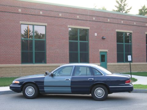 1991 Ford LTD Crown Victoria - A police car with a dark blue rear and a lighter blue front is parked outside a brick building with large windows.