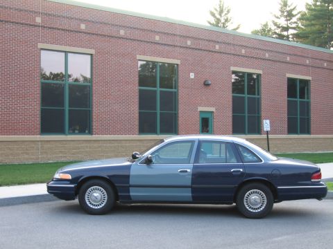 1991 Ford LTD Crown Victoria - A side view of a two-tone police car parked in front of a brick building with large windows.
