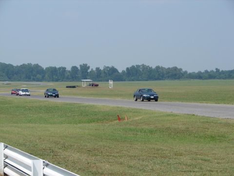 1992 Ford Taurus - A dark sedan drives on a racetrack with other vehicles in the background under a hazy sky.