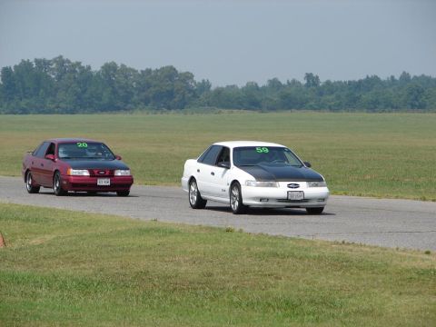 1992 Ford Taurus - Two Ford Taurus sedans compete on a race track, one white with a black hood and number 59, the other red with number 20.