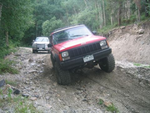 1996 Jeep Cherokee - Two SUVs navigate a rough, wooded trail. The lead Jeep Cherokee is red and appears to be a 1996 model.