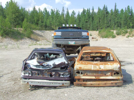1988 Ford Bronco - Head-on view of a dark teal Ford Bronco with roof-mounted driving lights and a green vanity plate, parked behind two stripped car bodies in…