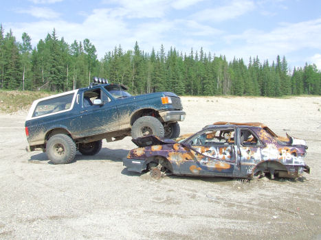 1988 Ford Bronco - Dark teal Ford Bronco with a white top and roof lights parked with its front tires up on the crushed, burned-out shell of a rusted sedan in…