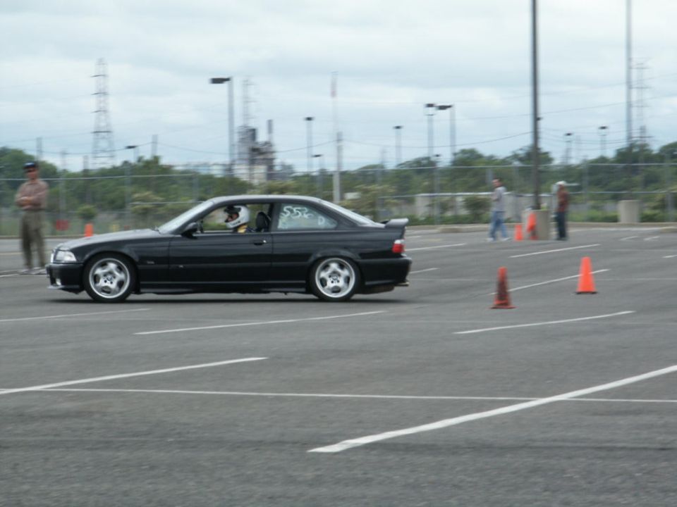 Black coupe driving on asphalt with orange cones. Driver wears a helmet. "S52" is visible on the car