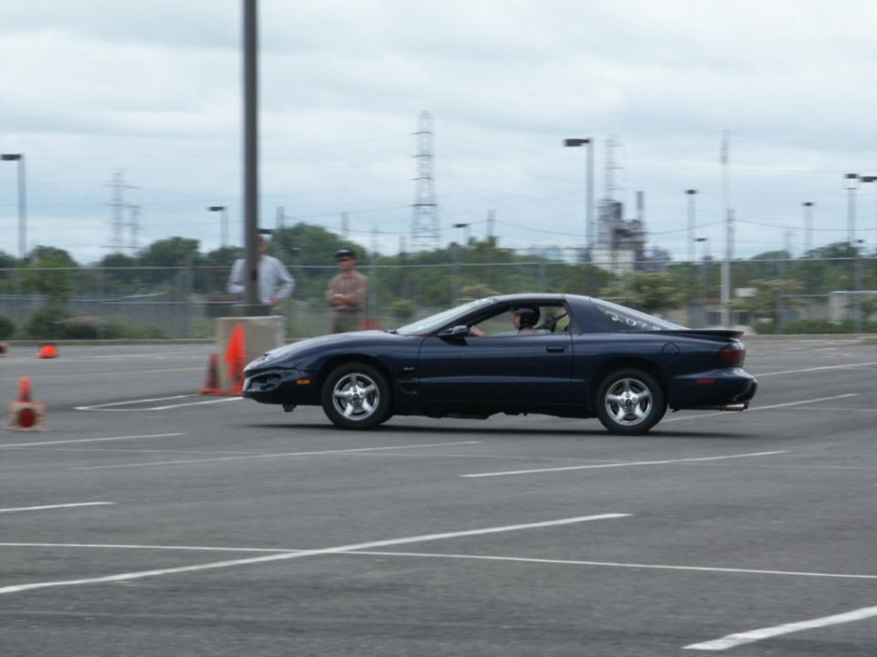 Dark blue coupe with silver wheels driving on a paved lot with orange cones. Text on rear quarter panel reads '207'