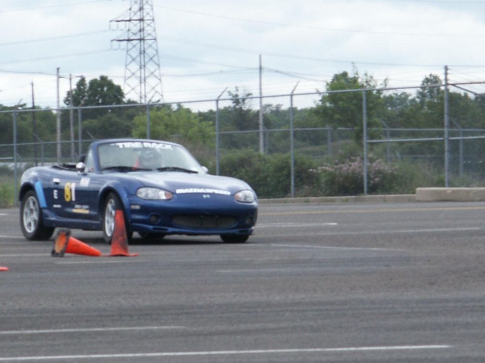 Blue convertible sports car with number 81 decal and 'Tire Rack' text on windshield, seen from a 3/4 front angle on a paved surface with orange cones