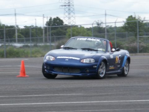 2001 Mercury Grand Marquis - A blue Mazda Miata convertible, identified by 'Mazdaspeed' and 'Tire Rack' branding, navigates a track with an orange cone nearby.