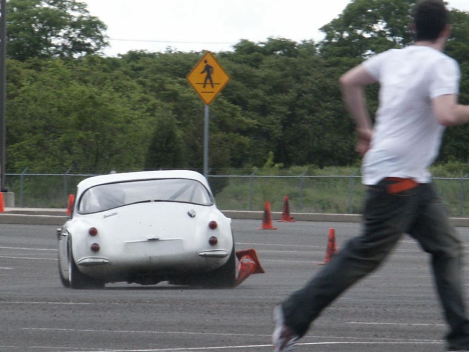 White vintage car with round taillights and a license plate area, seen from the rear at an auto-cross event with orange cones