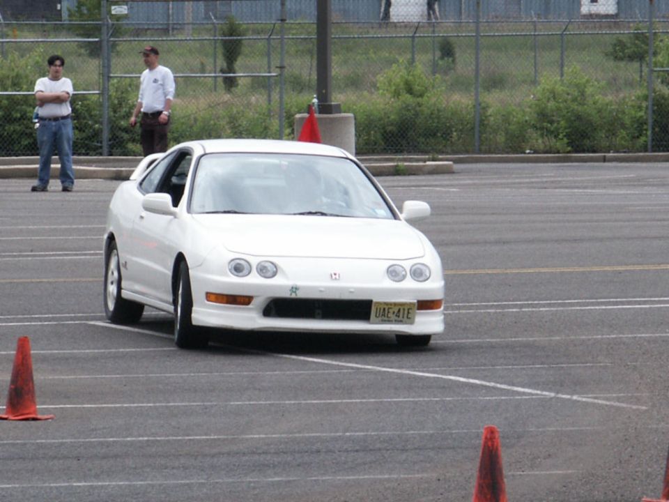 White coupe with pop-up headlights and a rear spoiler, driving on a paved lot