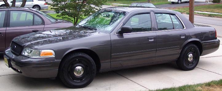 Dark gray Ford Crown Victoria sedan with black steel wheels and A-pillar spotlight parked in a driveway