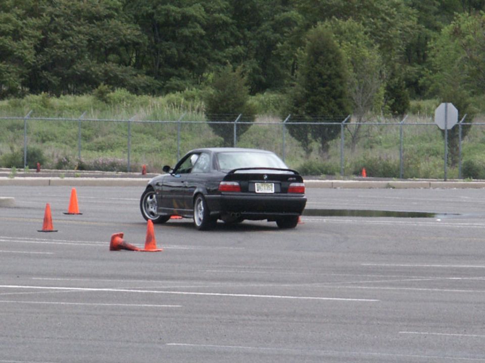 Black coupe with a spoiler and a license plate, seen from the rear on a paved lot with orange cones