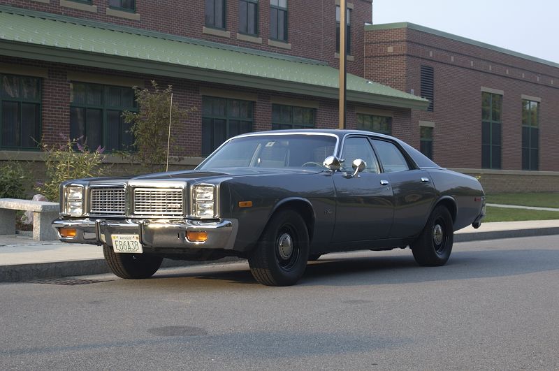 Charcoal 1970s Dodge four-door sedan, front-left three-quarter view, parked by brick building.