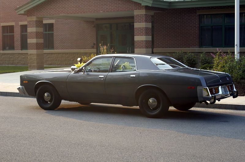 Dark gray 1970s four-door sedan, rear three-quarter view, parked by brick building