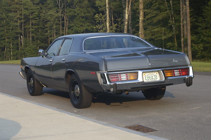 Rear three-quarter view of a gray 1970s Dodge coupe on pavement near trees.