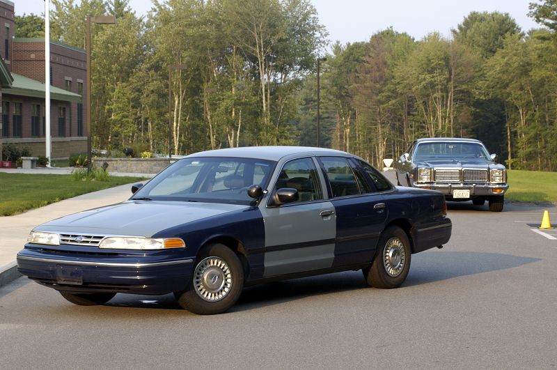 Dark-blue Ford Crown Victoria sedan with a primered fender; a vintage sedan behind.