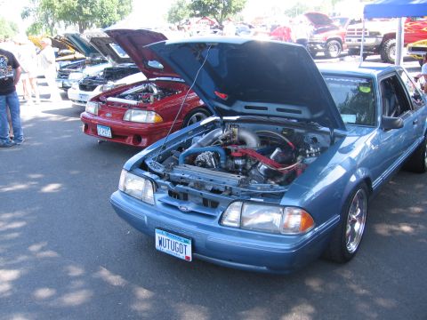 1989 Ford Mustang - A blue 1989 Ford Mustang is displayed with its hood open, revealing a modified engine bay, alongside other vehicles.