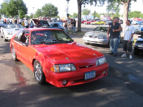 1989 Ford Mustang - A red 1989 Ford Mustang is the main subject at a car meet, with other vehicles and people in the background.