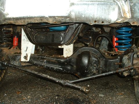 1985 Ford Bronco - Front-on view under a lifted Ford 4x4 showing the Dana solid front axle with its cast pumpkin, tie rod and drag link, blue lift coil spring…