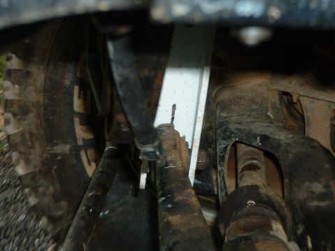 1985 Ford Bronco - Tight underbody shot of a lifted 4x4 front end, looking past a mud tire at the steering drag link, tie rod and radius arm bolted to a dirt-…