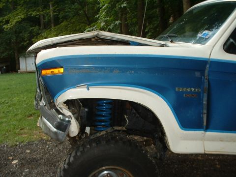 1985 Ford Bronco - Close-up of the driver's front corner of a blue and white two-tone Ford Bronco XLT with crash damage: the hood is buckled and peeled upward…