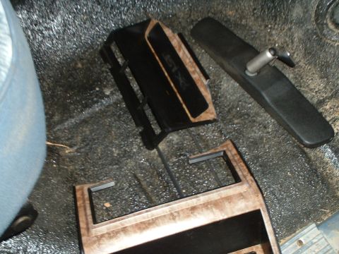 1985 Ford Bronco - Removed interior trim pieces laid on a dark carpeted vehicle floor, including a woodgrain-finished radio/center dash bezel, a black plastic…