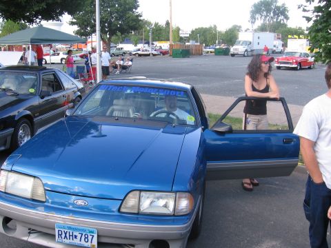 1989 Ford Mustang - A blue Ford Mustang is parked with its doors open, while people stand around it at an outdoor event.