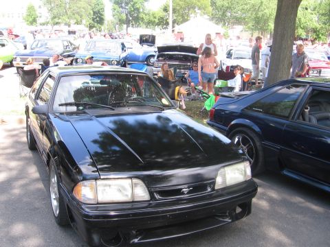 1989 Ford Mustang - A black Ford Mustang is parked at an outdoor car show with other vehicles and people around.
