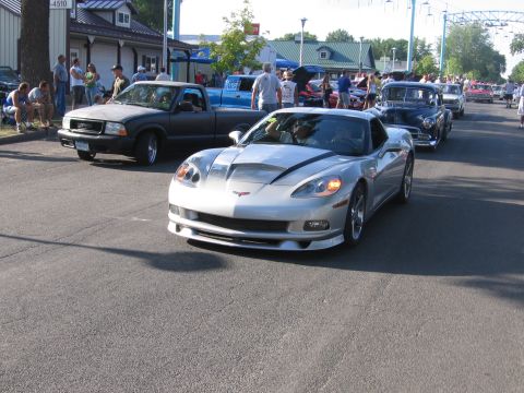 1989 Ford Mustang - A silver Chevrolet Corvette C6 leads a procession of vehicles down a street on a sunny day.