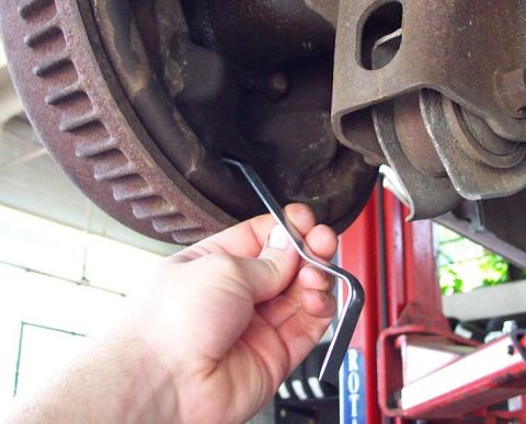 1985 Ford LTD Crown Victoria - A hand holds a bent brake pad anti-rattle clip beneath a rusty vented brake rotor and caliper bracket on a car raised on a red shop lift. T…