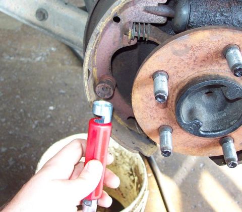 1985 Ford LTD Crown Victoria - A hand holding a red-handled brake spoon/adjusting tool up to a rear drum brake with the drum removed, showing the rusty five-lug axle flan…
