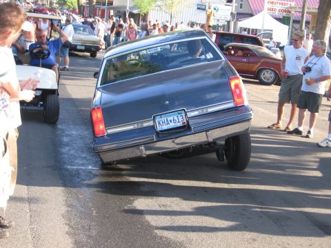 1989 Ford Mustang - A dark blue sedan is parked at an angle on a street with people around.