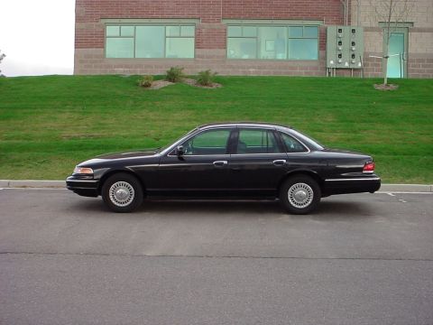 1998 Ford Crown Victoria - Side profile of a black 1998 Ford Crown Victoria parked on asphalt in front of a grassy hill and brick building.