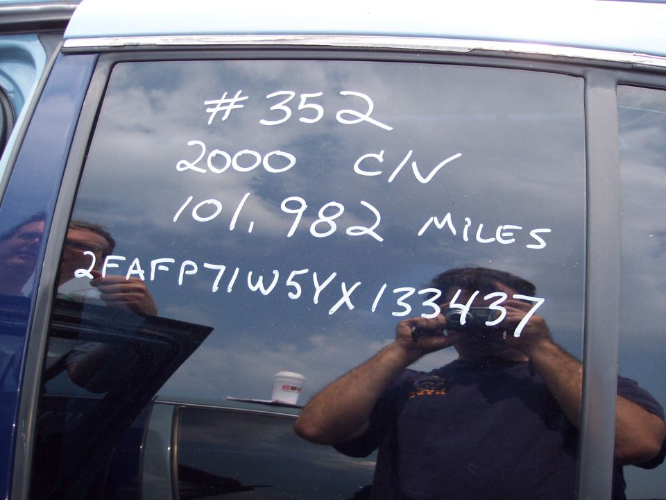 Blue car window with white writing: # 352, 2000 C/V, 101,982 Miles, 2FAFP71W5YX133437. Reflections of people and cameras