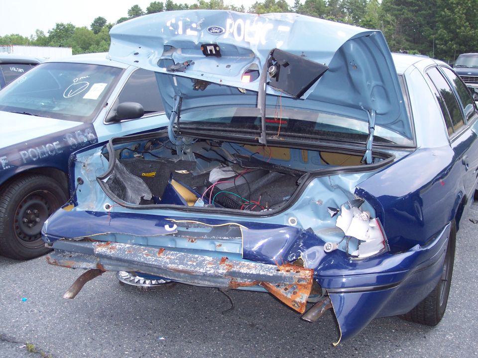 Damaged blue police car rear with trunk open, showing exposed wires and bent metal