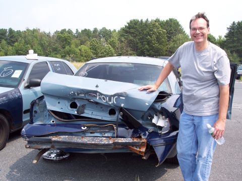 1991 Ford LTD Crown Victoria - A man stands next to a severely damaged police car, with another damaged police car visible behind it.