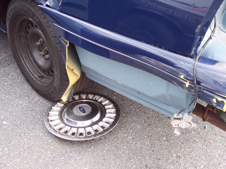 Ford hubcap lying on asphalt near a damaged blue car fender and exposed yellow inner fender liner