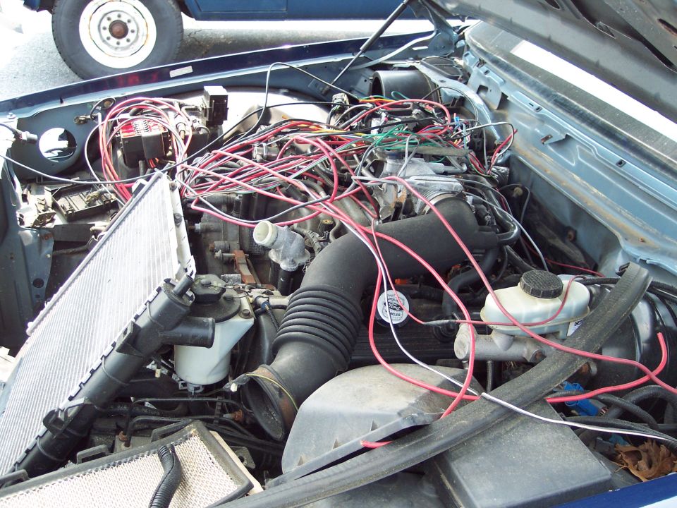 Engine bay with tangled wires and a detached radiator. Another vehicle's wheel is visible