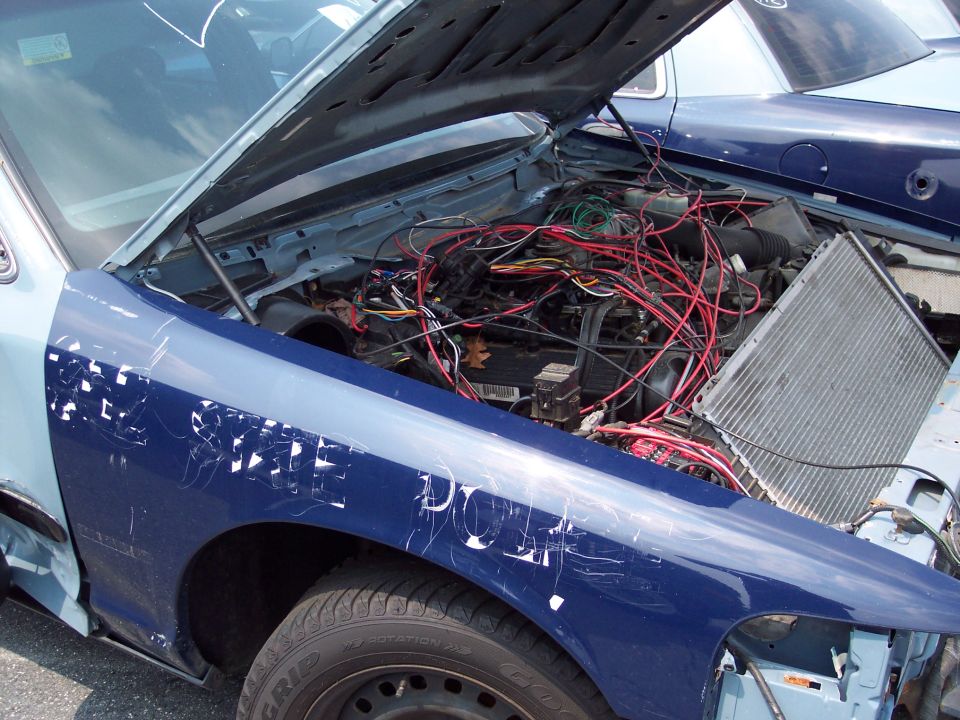 Engine bay of a blue and light blue car with hood open, showing tangled wiring and a detached radiator. Another blue car is visible behind it
