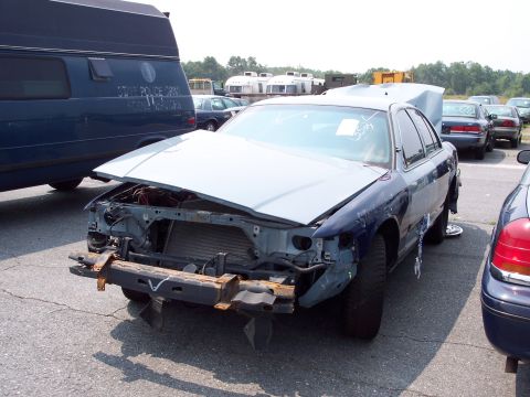 1991 Ford LTD Crown Victoria - A damaged blue sedan sits in a lot with its hood open and front end dismantled.