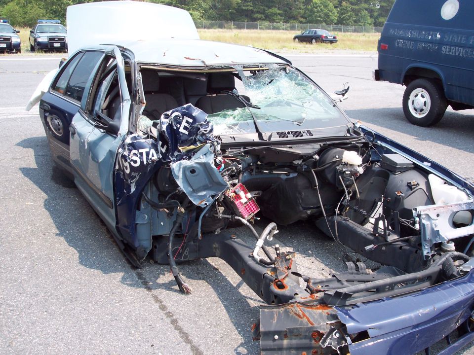 Front end of a wrecked blue police car with 'STATE POLICE' visible on the side. Hood is up, windshield is shattered, and the front is heavily damaged