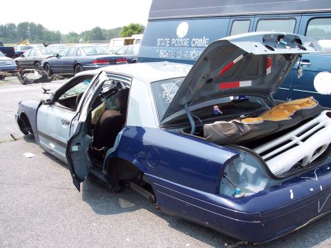 1991 Ford LTD Crown Victoria - A heavily damaged 1991 Ford Crown Victoria LTD police car sits in a lot with its trunk and doors open, missing wheels.