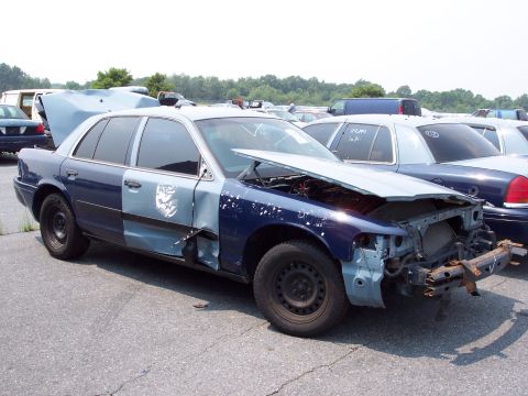 1991 Ford LTD Crown Victoria - A damaged police car sits in a junkyard with its hood open and front end dismantled.