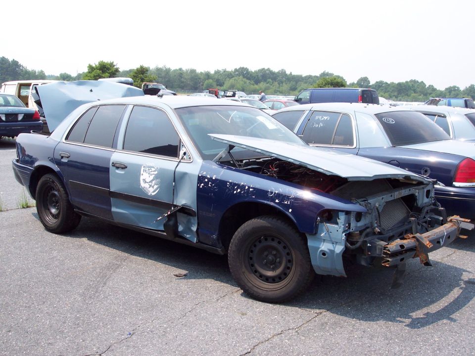 Damaged police car with hood open, missing front bumper and grille. Two-tone blue and light blue paint job