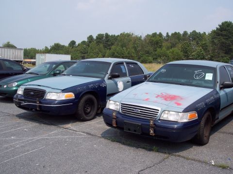 1991 Ford LTD Crown Victoria - Two police cruisers are parked side-by-side in an outdoor lot under a clear sky.